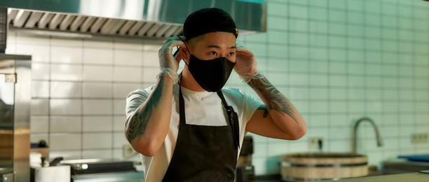 masked asian chef during quarantine days. young worker of a restaurant standing in a large kitchen. quarantine cook room concept