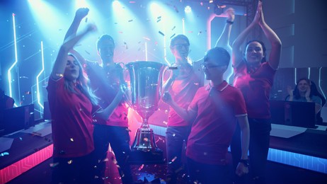 diverse esport team winner of the video games tournament celebrates victory cheering and holding trophy in big championship arena. cyber gaming event with gamers and fans.