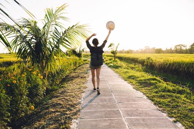 back view of carefree hipster girl with travel backpack walking at path way feeling freedom during summer vacations, happy woman raising straw hat in hand enjoying youthful touristic lifestyle