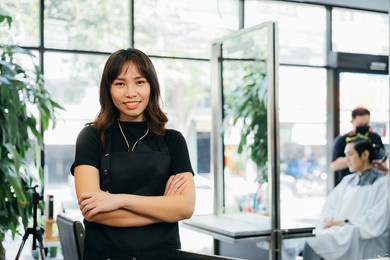 portrait of happy successful young asian salon owner standing wearing apron with folded hands smiling while looking at camera