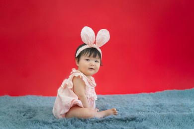 baby in easter bunny hairband. asian happy baby smiling and sitting on red background. cute 6 months baby start sitting with copy space as easter concept, baby or kid department in hospital.