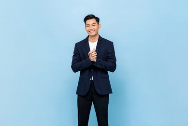 smiling young handsome southeast asian man clapping hands impressively in light blue studio isolated background
