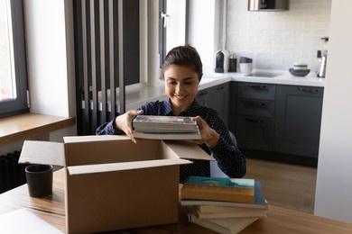 smiling indian woman unpacking parcel, holding books, sitting at table, satisfied customer received online store order, delivery service, happy female packing belongings, preparing for relocation