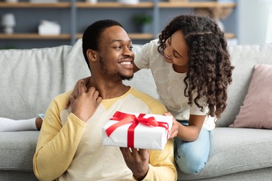 adorable black girl greeting her smiling young father, hugging him and giving gift box, living room interior, copy space. happy african american daddy and daughter celebrating fathers day at home