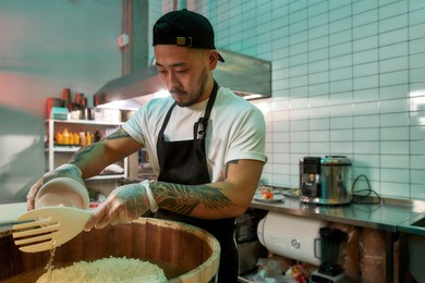 young male chef cook looking busy while making sushi rice in a wooden barrel in the kitchen of japanese restaurant. asian cuisine concept. horizontal shot