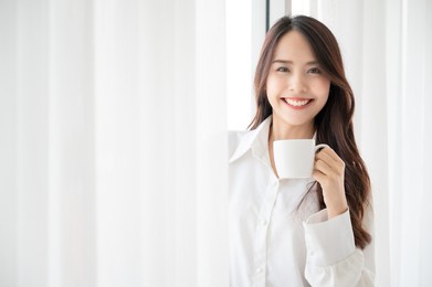 young asian woman standing beside window and holding mug in bedroom at home, she drinking milk after wake up in the morning