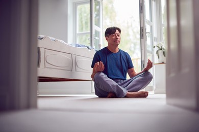 mature asian man in pyjamas sitting on bedroom floor meditating in yoga pose