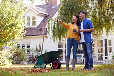 mature asian couple taking a break with hot drinks whilst tidying garden with rake and barrow