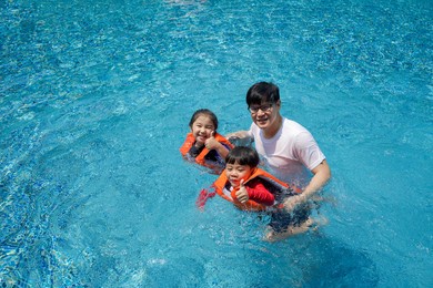 asian father teaches his son and daughter to swim in the pool. the older sister and younger brother wearing orange life jacket, smiling with thumbs up.
