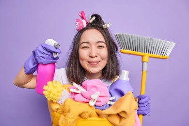isolated shot of positive brunette asian housewife holds detergent for cleaning holds broom poses near laundry basket does disinfection of house isolated over purplebackground. cheerful housekeeper