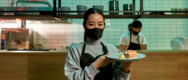 portrait of young waitress wearing protective mask looking at camera and holding a plate with sushi rolls. japanese cuisine in restaurant. asian food concept. web banner