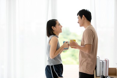 portrait shot of cute smiling young asian lover couple standing near the windows in the living room in morning, holding and drinking cup of coffee while looking at each other eyes and talking with fun