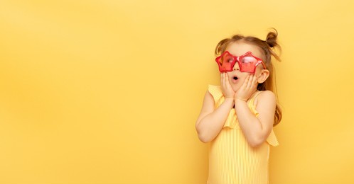 little child girl in striped swimsuit and red funny summer sunglasses surprised expression looks at camera posing on yellow background, studio portrait.advertising of children's products and sale