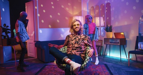happy young caucasian stylish guy sitting in chair in living room in neon disco ball light, looking at camera and smiling while multi-ethnic friends dancing and moving behind having fun at party time