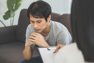 young asian man, male has mental symptoms which must have been therapy and stress, sitting on couch to consult to psychologist during the session  taking notes to find out how to treat the therapist.
