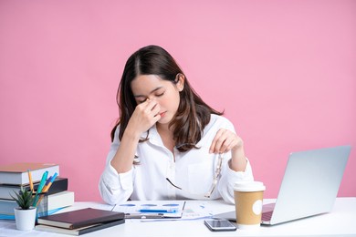 young asian woman are stressed and tired from work. she was holding glasses at a white desk with a laptop in the office. isolated on a pastel pink background.