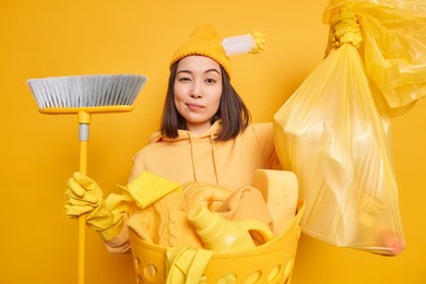 photo of serious asian woman wears hat rubber latex gloves collects rubbish in house holds big polythene bag full of wastes broom for sweeping floor busy doing house chores isolated on yellow wall