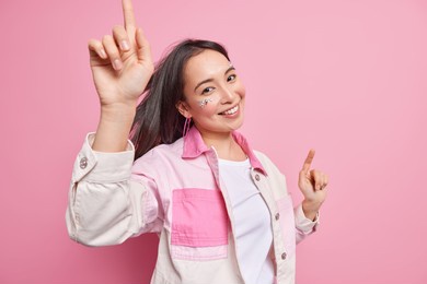 positive lovely asian woman raises arms while dances to music expresses joy and happiness feels relaxed catches every bit of song poses half turned against pink background. happy emotions concept