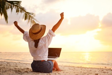 work from anywhere. rear view of young woman, female freelancer in straw hat working on laptop, keeping arms raised and cellebrating success while sitting on the tropical sandy beach at sunset