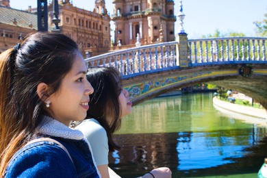 asian women tourist. they are leaning on the railing looking at the back of the square. in the background a tower and a bridge over a river. they are in seville. spain