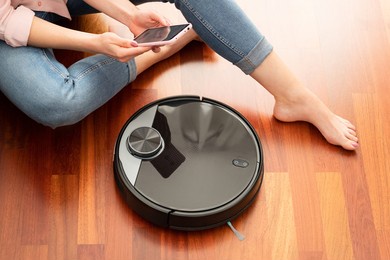 faceless middle section of young woman using automatic vacuum cleaner to clean the floor, controlling smart machine housework robot with smart phone