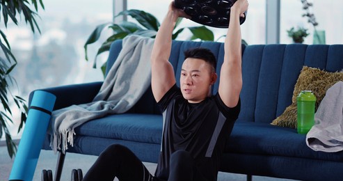 handsome strong asian young man engaged in heavy sports bodybuilding lifting barbell disc practicing strength endurance working out in living room.