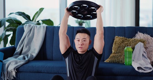 chinese young strong sportsman leaning to sofa lifting abs with barbel heavy disc practicing effort training at home. workout in apartment.