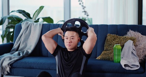 handsome strong asian young man engaged in heavy sports bodybuilding lifting barbell disc practicing strength endurance working out in living room.
