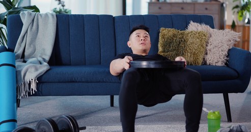 chinese young strong sportsman leaning to sofa lifting abs with barbel heavy disc practicing effort training at home. workout in apartment.