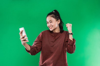 portrait beautiful woman holding and look handphone with happiness expression while raising one hand and making a fist, isolated on green background