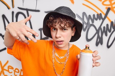 funky self confident hipster guy makes yo gesture enjoys cool music holds aerosol spray creats graffiti wall on public space wears hat casual t shirt. urban guy makes live murales paint by himself