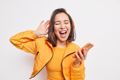 overjoyed pretty asian teenage girl got carried away with music holds smartphone enjoys favorite playlist via stereo headphones has relaxed carefree expression sings along wears orange jacket