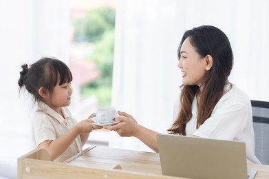 cute happy little daughter give coffee to her smiling mother while mom works on computer laptop at home on holiday