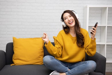 portrait of a happy young asian woman listening music with wireless headphones from music application on mobile smart phone in living room at condominium