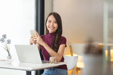 young asian woman using phone and laptop in coffee shop and looking at camera.