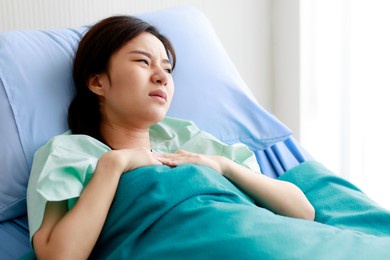 asian beautiful young female patient lying on bed in hospital and touching her chest with pain while being heart disease. medical treatment and health concept