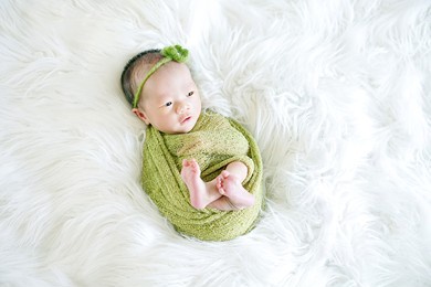 opened eyes newborn wrapped in a green cloth with a green flower headband showing little feet lying on a soft white flokati
