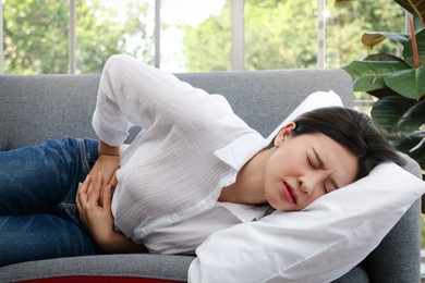 a beautiful asian woman has a stomachache lying on the sofa in her room at home. hospital concept woman sickness warning signs of diseases or abdominal pain before menstruation.