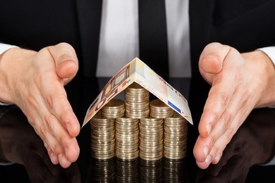 midsection of young businessman protecting house made of currency at desk