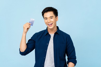 waist up portrait of happy smiling young handsome asian man holding credit card in isolated light blue background