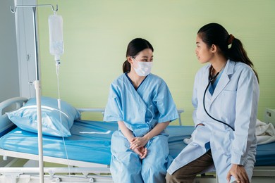 young asian female doctor with patient wearing surgical face mask sitting on hospital bed talking to each other and talking routine status update