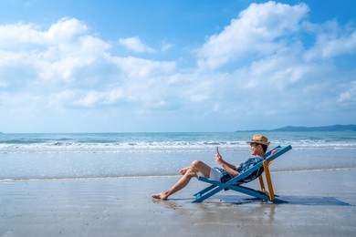 young asian man resting on sunbed on tropical beach. happy guy sit on beach chair by the sea using smartphone for selfie or video call. handsome male enjoy beach outdoor lifestyle on summer vacation