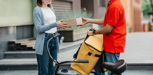 courier service worker on bike delivers food, parcel to customer, online ordering city shipping. young man in uniform with yellow thermal bag gives box to woman to client, side view, panorama, cropped