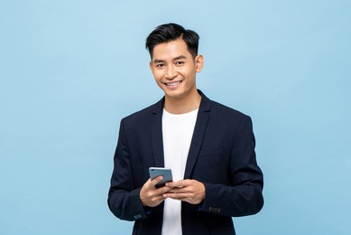 waist up portrait of young smiling handsome asian man in semi formal suit using mobile phone in light blue isolated studio background