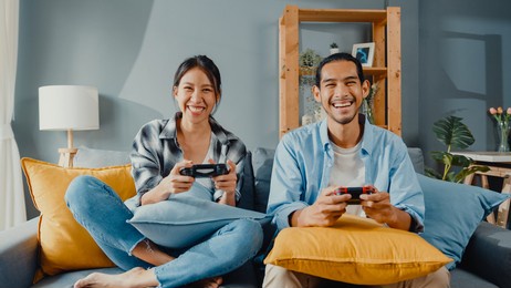 happy asia young couple man and woman sit on couch use joystick controller play video game spend fun time together in living room. asian married couple family lifestyle, couple stay at home concept.
