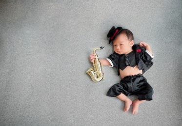 top of adorable baby playing music by gold saxophone wearing black suite and tuxedo hat decorated with red rose on shirt. newborn lying on grey background.