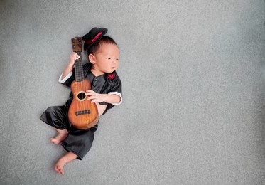 top of cute newborn baby wearing suites with tuxedo hat and red rose on suit. the child playing ukulele guitar lying on grey backgroud.
