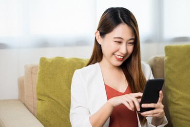 young asian business woman sitting on sofa in living room at home using smartphone and smiling.