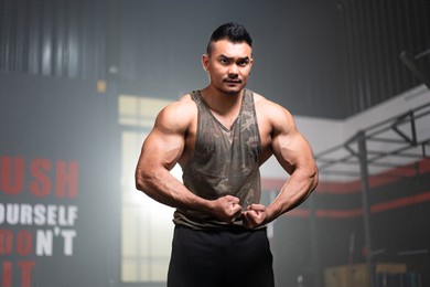 young asian muscular male fitness trainer in a tank top standing with hold his arms down, flexing arm and shoulder muscles while looking forward with a serious face with a blurred background of gym.