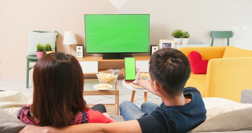 asian young couple sit on couch in living room watching tv and looking at smartphone with a green screen 